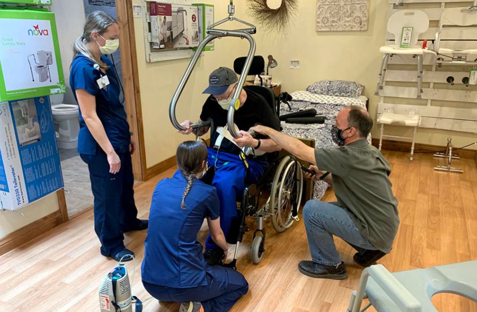 an elder man in a wheelchair is assisted by OT's and an Accessible Systems specialist as he is fitted for the SureHands ceiling lift system