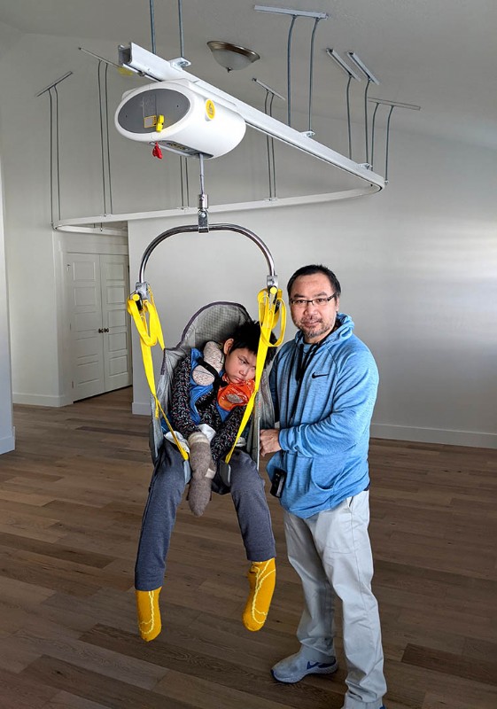 A father stand next to his disabled son that is suspended by a SureHands ceiling lift